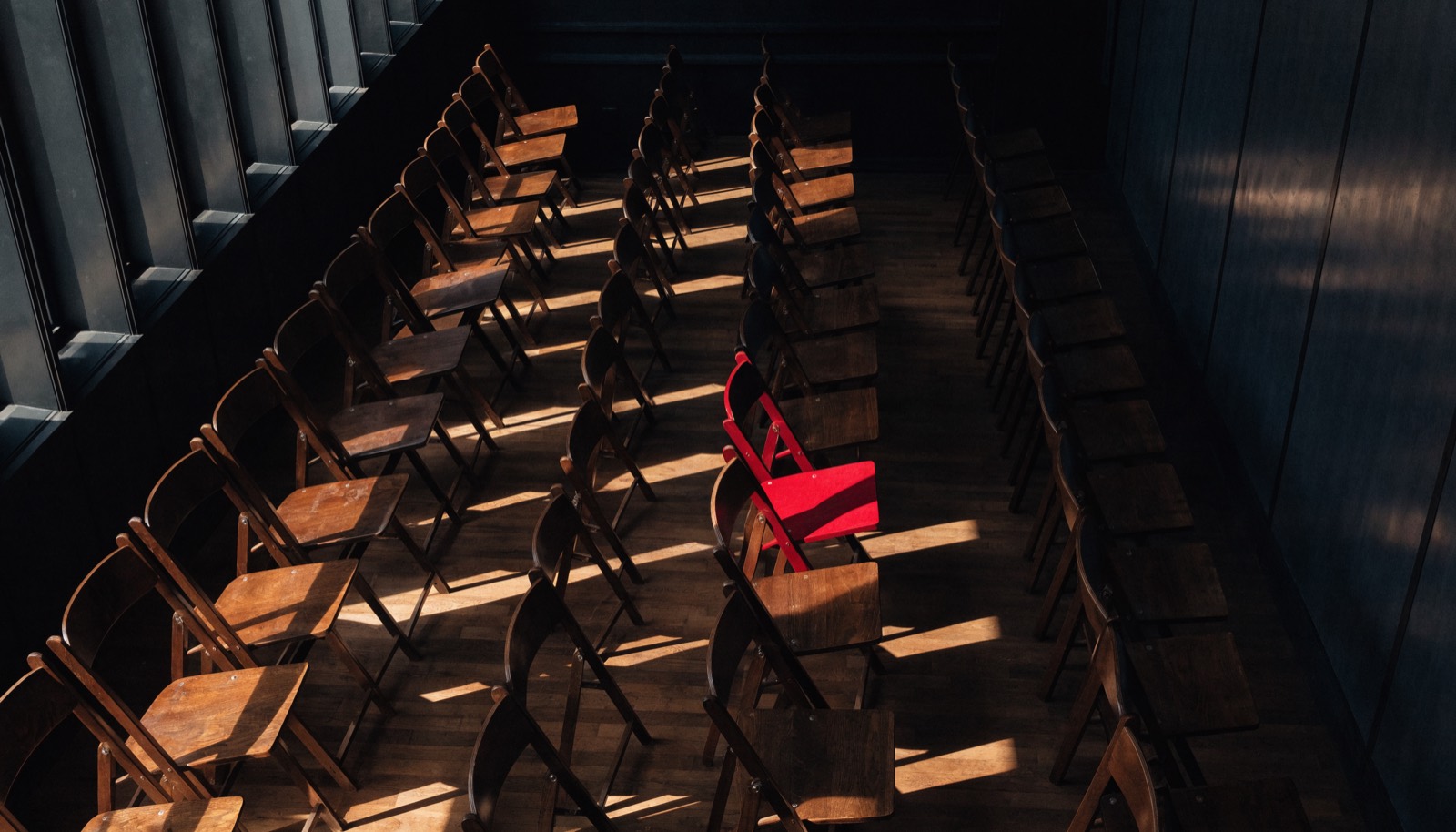 Rows of empty wooden folding chairs in a large hall, lit from tall windows, one chair crimson.
