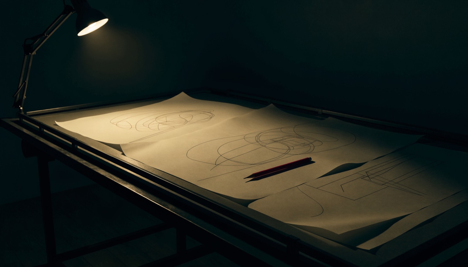 A drafting table lit by one hard lamp in a dark room, with large pale technical drawings and a crimson pencil.